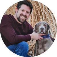 A man sitting outdoors with a gray dog in front of tall grass.